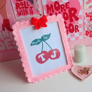 以下が含まれることがあります： 「Made by TJ」と書かれたさくらんぼのグラフィックと小さな赤いリボンが付いた、ピンク色の手編みのフレーム。フレームは白い表面にあり、ハート型の皿とキャンドルの山があります。背景には装飾的なポスターがあります。