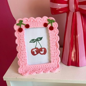 May include: A pink, crocheted picture frame with a scalloped edge. The frame has cherry accents and a picture with a cherry graphic and the words "Made by". A red and pink striped decorative bow is in the background.
