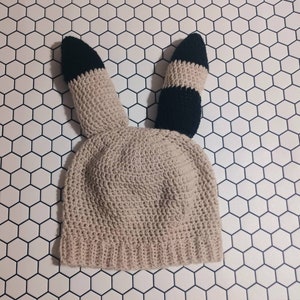 May include: A beige crocheted hat with black and white striped ears. The hat is on a white background with a hexagonal pattern.