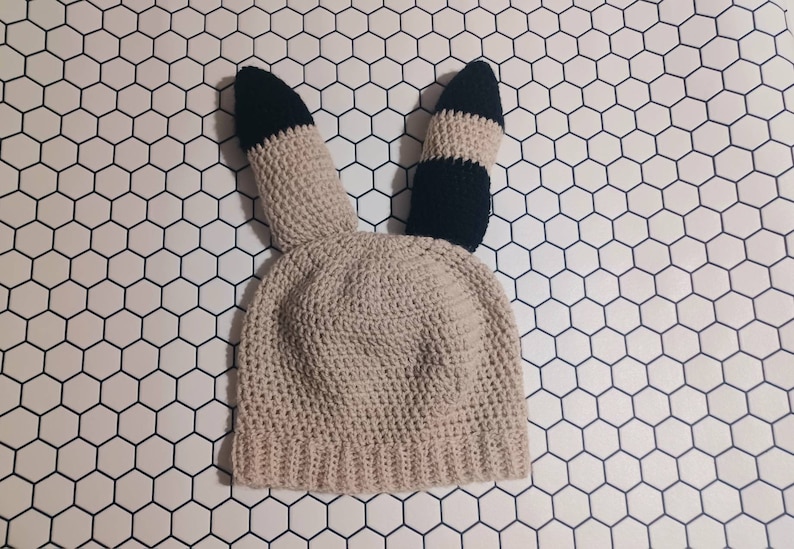 May include: A beige crocheted hat with black and white striped ears. The hat is on a white background with a hexagonal pattern.
