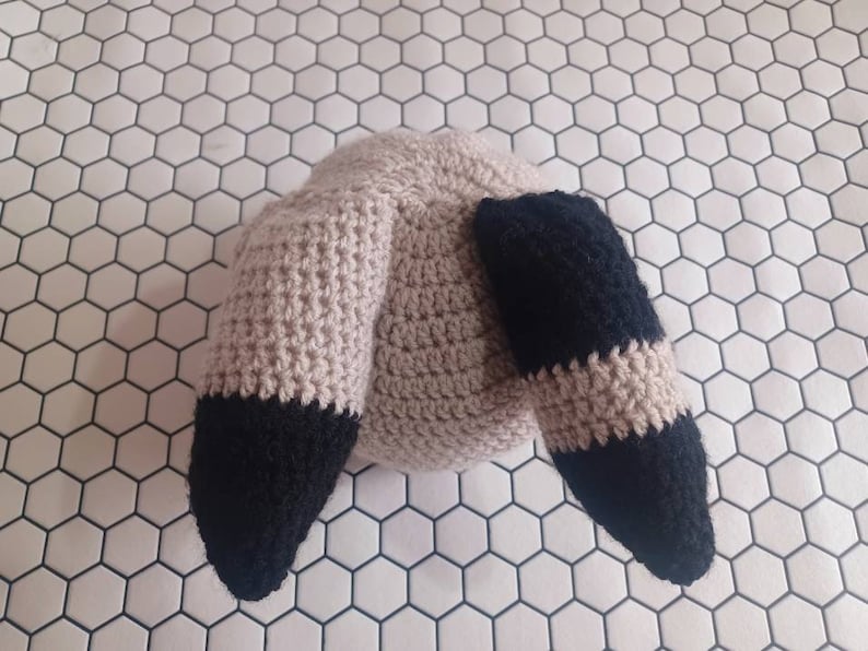 May include: A crocheted stuffed animal with a light brown body and two black ears. The ears are pointed and have a black and brown striped pattern.