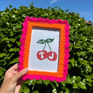 May include: A handmade, rectangular picture frame with a bright pink scalloped edge, an orange inner border, and a white interior. The frame holds a print with a cherry design and the text "Made by TJ".