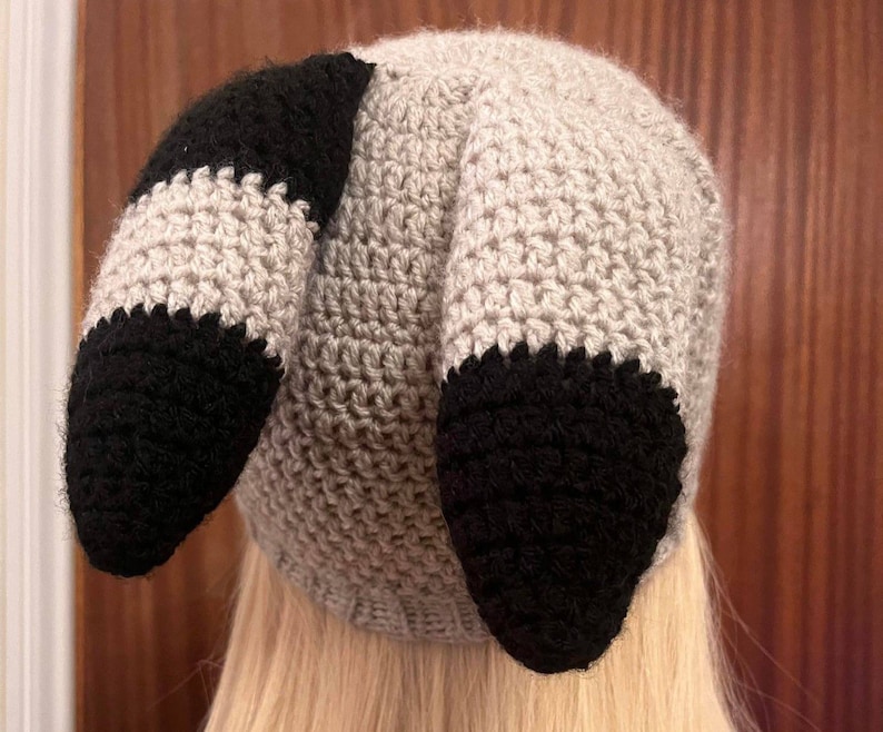 May include: A gray and black crocheted hat with two long, floppy ears. The hat is made with a textured stitch and has a black and gray color scheme.