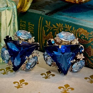 May include: A pair of vintage-style earrings with a triangular, deep blue center stone and smaller light blue accent stones. The earrings are set in a gold-toned metal and rest on a patterned surface. A gold decorative box and a glass vase with white flowers are in the background.