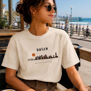 Könnte beinhalten: Ein cremefarbenes Kurzarm-T-Shirt mit einer Grafik der Skyline von Busan, einer Brücke und einer Sonne. Der Text "BUSAN" und "where skyscrapers meet the sea" ist in schwarzen Buchstaben gedruckt. Der Stoff hat eine dezent gesprenkelte Textur.