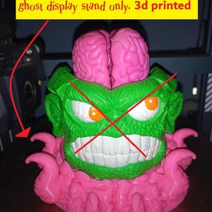 May include: A 3D printed display stand for The Real Ghostbusters Brain Blaster ghost. The object features a green monster head with orange eyes and white teeth, topped with a pink brain, and surrounded by pink tentacles at the base.