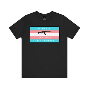 Puede incluir: Camiseta negra con una bandera del orgullo transgénero y el texto "BE AN ALLY ARM YOUR TRANS FRIENDS" con una silueta de un rifle de asalto en la bandera.