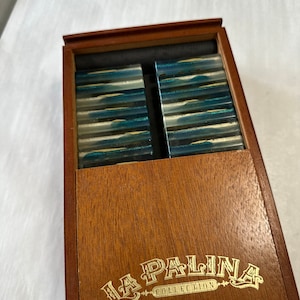 Puede incluir: Una caja de madera con una inscripción dorada que dice "La Palina Collection Mr. Sam". La caja contiene dos pilas de cigarros azules y blancos.