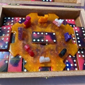 Puede incluir: Una caja de madera con una incrustación de resina dorada y negra y un juego de dominó. Los dominós están dispuestos en círculo alrededor de la incrustación. También hay varias pequeñas figuritas de colores en la incrustación.