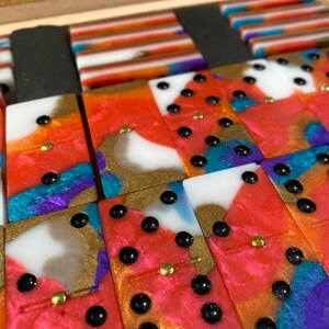 Puede incluir: Un juego de dominó de colores con puntos negros y detalles dorados. Los dominós están hechos de un material translúcido con un patrón de remolino de rosa, azul, morado y dorado.