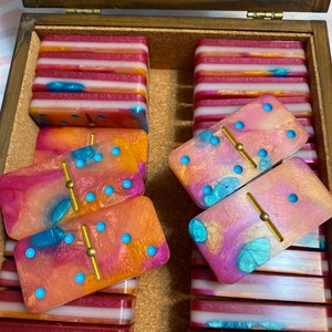 Puede incluir: Una caja de madera llena de dominós coloridos. Los dominós tienen un diseño marmoleado rosa, naranja y azul con puntos dorados y una línea divisoria dorada. Los dominós están apilados en la caja.