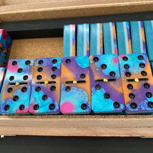 Puede incluir: Un juego de seis dominós coloridos con un patrón de remolino azul, morado y naranja. Cada dominó tiene una línea negra y puntos dorados.