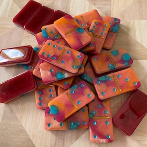 Puede incluir: Un juego de dominó con un diseño abstracto y colorido. Los dominós están hechos de resina y tienen un acabado brillante. Los dominós son de varios colores, incluyendo rojo, naranja, amarillo y azul. Los dominós están dispuestos en una pila sobre una superficie de madera.