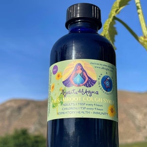 Op de afbeelding: Een donkerblauwe glazen fles met een zwarte dop, met het label "Mjercita del Agua Balsamroot Cough Syrup". Het label heeft een kleurrijke illustratie van een vrouw die een kopje water vasthoudt. De fles staat op een oppervlak met een wazige achtergrond van een berg en de lucht.