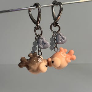 May include: Two keychains featuring small, sculpted fish charms hanging from silver-toned heart-shaped lobster clasps. One fish is brown with white spots, the other is peach-colored. Each includes a small lavender heart bead and light blue accents.
