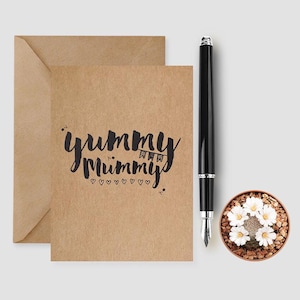May include: A kraft paper greeting card and envelope with the words "yummy mummy" in black script. A black fountain pen and a small potted cactus with white flowers are also present.