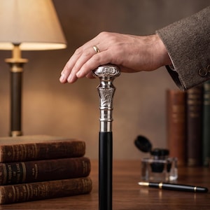 Op de afbeelding: Decoratieve wandelstok met een zilveren handvat en een zwarte schacht. De zilveren handgreep heeft ingewikkelde patronen. De stok wordt vastgehouden door een persoon die een bruine tweedjas draagt. Boeken en een bureaulamp op de achtergrond.