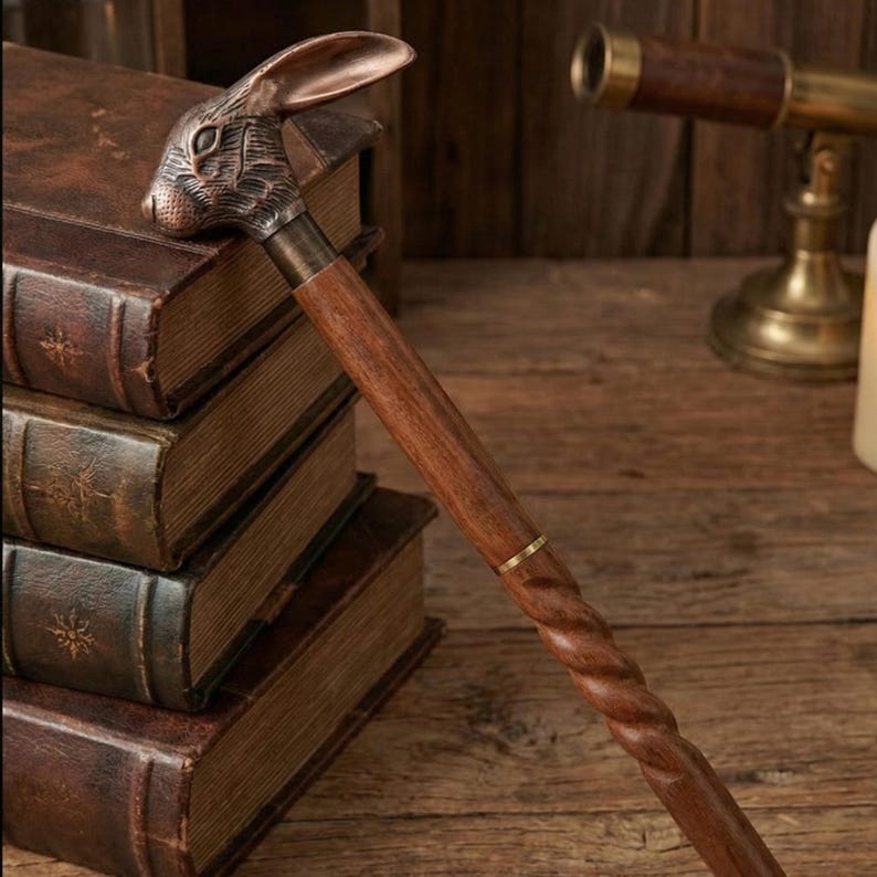 Puede incluir: Bast&oacute;n decorativo con un mango en forma de cabeza de conejo. El mango es de color bronce y el eje es de madera oscura con un dise&ntilde;o retorcido. El bast&oacute;n est&aacute; apoyado contra una pila de libros antiguos.