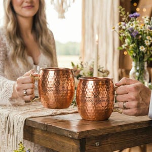 Puede incluir: Dos tazas de cobre con textura martillada y asas doradas se muestran sobre una mesa de madera. Cada taza tiene el texto "HANNAH & ANDREW Est. 2024" con un diseño de corazón. Las tazas son parte de un tema de boda.