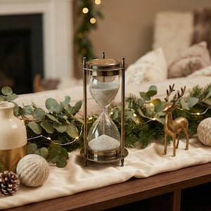 Puede incluir: Un reloj de arena de estilo antiguo con marco de latón y cristal transparente. La arena del interior es de color gris claro. El reloj de arena está sobre una mesa de madera con objetos decorativos, incluyendo una figura de ciervo dorada y un jarrón blanco.