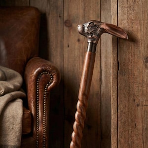 Può includere: Bastone da passeggio decorativo con manico a forma di testa di coniglio in bronzo e fusto in legno intrecciato. Il bastone è appoggiato a una parete di legno, con una poltrona in pelle sullo sfondo.