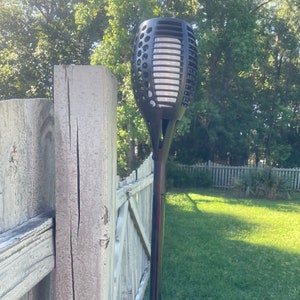 May include: A black solar powered torch light mounted on a wooden fence post. The light is shaped like a flame and has a white plastic diffuser. The light is mounted on a black metal pole.