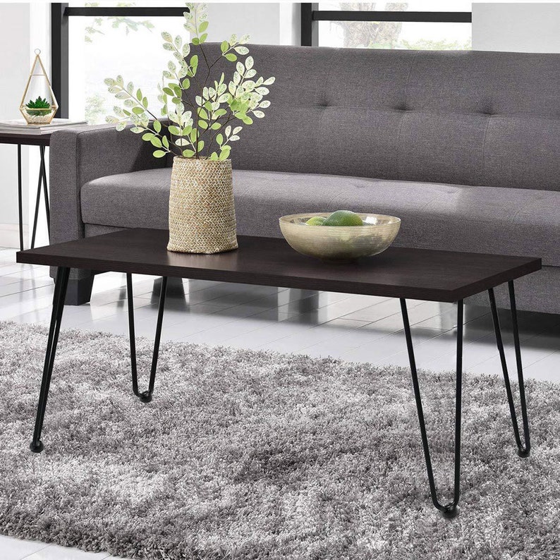 Op de afbeelding: Een donkerbruine houten salontafel met zwarte haarspeldpoten. De tafel staat op een grijs tapijt voor een grijze bank. Een vaas met groene planten en een kom met fruit staan op de tafel.
