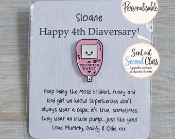 May include: A pink enamel pin shaped like an insulin pump with a smiling face and the text YOU'RE TOO SWEET, attached to a white card featuring the text Sloane Happy 4th Diaversary and a message about superheroes wearing insulin pumps.