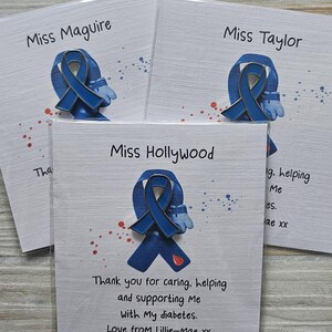 May include: Three blue awareness ribbon pins attached to white cards. Each card features a name at the top and text below the pin. The pin displays a blue ribbon with a small red droplet icon. The cards include printed messages of gratitude.