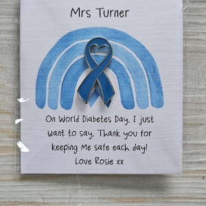 May include: A blue heart-shaped awareness ribbon pin attached to a white card featuring a blue watercolor rainbow. The card text reads: Mrs Turner, On World Diabetes Day, I just want to say, Thank you for keeping Me safe each day! Love Rosie xx.