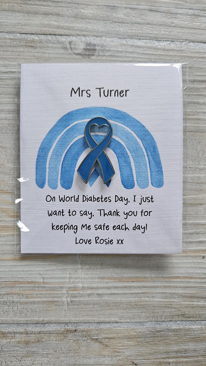 May include: A blue heart-shaped awareness ribbon pin attached to a white card featuring a blue watercolor rainbow. The card text reads: Mrs Turner, On World Diabetes Day, I just want to say, Thank you for keeping Me safe each day! Love Rosie xx.