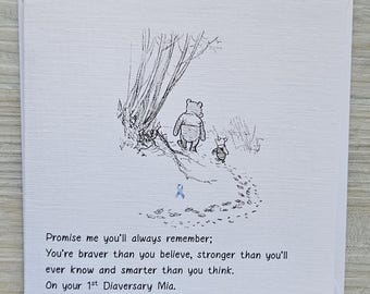 May include: A square white greeting card featuring a black ink illustration of Winnie the Pooh and Piglet walking along a path. A small blue ribbon is centered below them. The text reads: Promise me you'll always remember; You're braver than you believe, stronger than you'll ever know and smarter than you think. On your 1st Diaversary Mia.