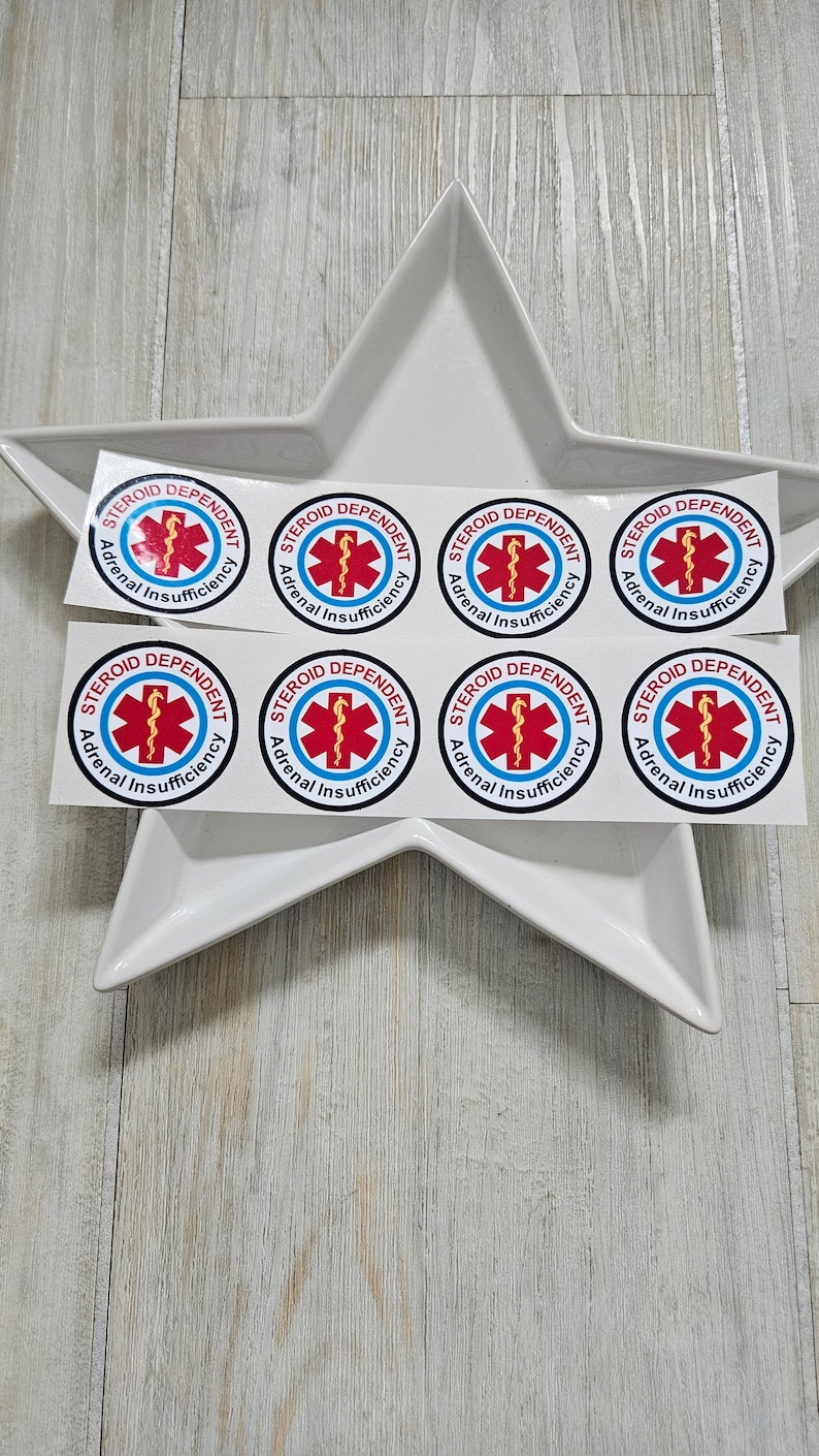 Pu&ograve; includere: Una collezione di adesivi rotondi con la scritta "STEROID DEPENDENT Adrenal Insufficiency". Presentano un simbolo medico rosso in un cerchio blu. Gli adesivi sono disposti su un piatto bianco a forma di stella, su sfondo di legno chiaro.