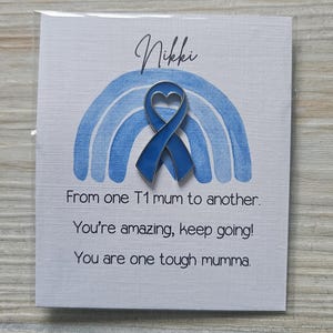 May include: A blue enamel heart-shaped awareness ribbon pin centered on a white card featuring a light blue watercolor rainbow. The card text reads: Nikki, From one T1 mum to another. You're amazing, keep going! You are one tough mumma.