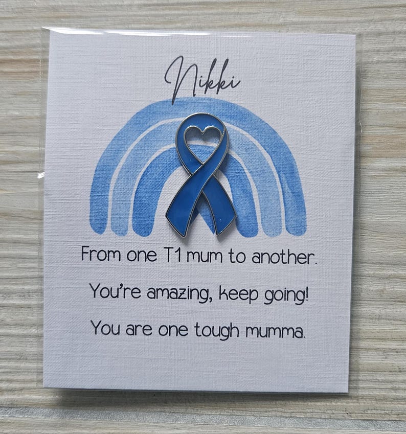 May include: A blue enamel heart-shaped awareness ribbon pin centered on a white card featuring a light blue watercolor rainbow. The card text reads: Nikki, From one T1 mum to another. You're amazing, keep going! You are one tough mumma.