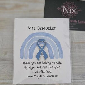 May include: A blue heart-shaped awareness ribbon pin attached to a white card featuring a watercolor blue rainbow illustration. The card text reads: Mrs DeMpster, Thank you for helping Me with My highs and lows this year. I will Miss You. Love Megan S (2024) xx.