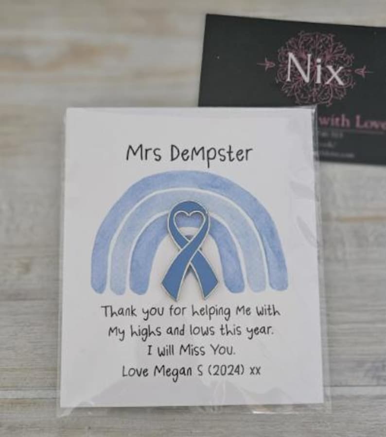 May include: A blue heart-shaped awareness ribbon pin attached to a white card featuring a watercolor blue rainbow illustration. The card text reads: Mrs DeMpster, Thank you for helping Me with My highs and lows this year. I will Miss You. Love Megan S (2024) xx.