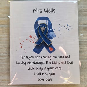 May include: A blue enamel awareness ribbon pin attached to a white card featuring the text Mrs Wells, a printed blue ribbon graphic, red paint splatters, and the message Thankyou for keeping Me safe and helping Me through the highs and lows.