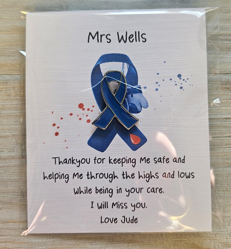 May include: A blue enamel awareness ribbon pin attached to a white card featuring the text Mrs Wells, a printed blue ribbon graphic, red paint splatters, and the message Thankyou for keeping Me safe and helping Me through the highs and lows.