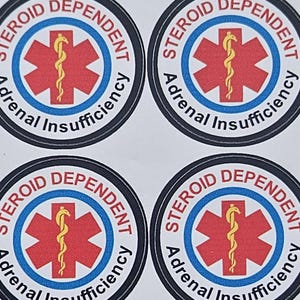 Pu&ograve; includere: Un foglio di otto adesivi circolari. Ogni adesivo presenta un simbolo medico rosso con un bastone giallo al centro, circondato da un cerchio blu. Il testo recita "STEROID DEPENDENT Adrenal Insufficiency" in un bordo nero.