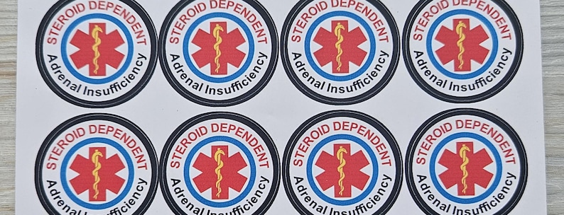 Pu&ograve; includere: Un foglio di otto adesivi circolari. Ogni adesivo presenta un simbolo medico rosso con un bastone giallo al centro, circondato da un cerchio blu. Il testo recita "STEROID DEPENDENT Adrenal Insufficiency" in un bordo nero.