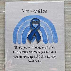May include: A blue enamel awareness ribbon pin with a heart-shaped cutout, centered on a white card featuring a blue watercolor rainbow. The card text reads: Mrs HaMilton, Thank you for always keeping Me safe throughout My highs and lows. you are aMazing and I will Miss you. FROM Teddy.
