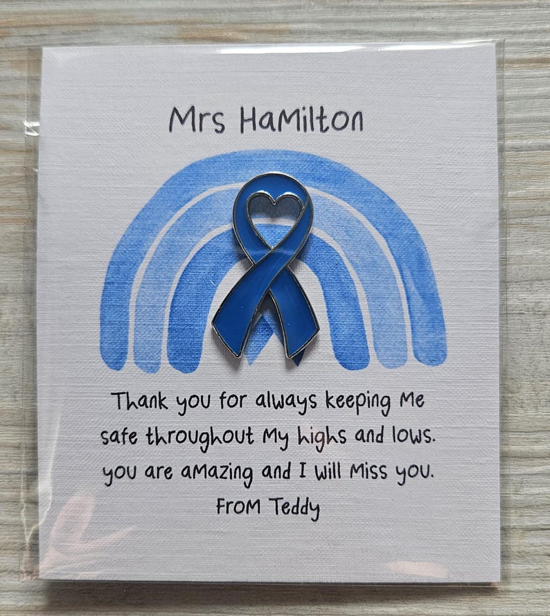 May include: A blue enamel awareness ribbon pin with a heart-shaped cutout, centered on a white card featuring a blue watercolor rainbow. The card text reads: Mrs HaMilton, Thank you for always keeping Me safe throughout My highs and lows. you are aMazing and I will Miss you. FROM Teddy.