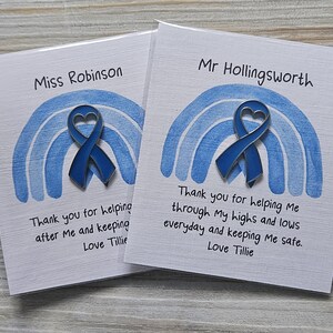 May include: Two white cards featuring a blue enamel heart-shaped ribbon pin centered over a blue painted rainbow graphic. The cards are personalized with names and printed text: Thank you for helping me through my highs and lows everyday and keeping me safe. Love Tillie.