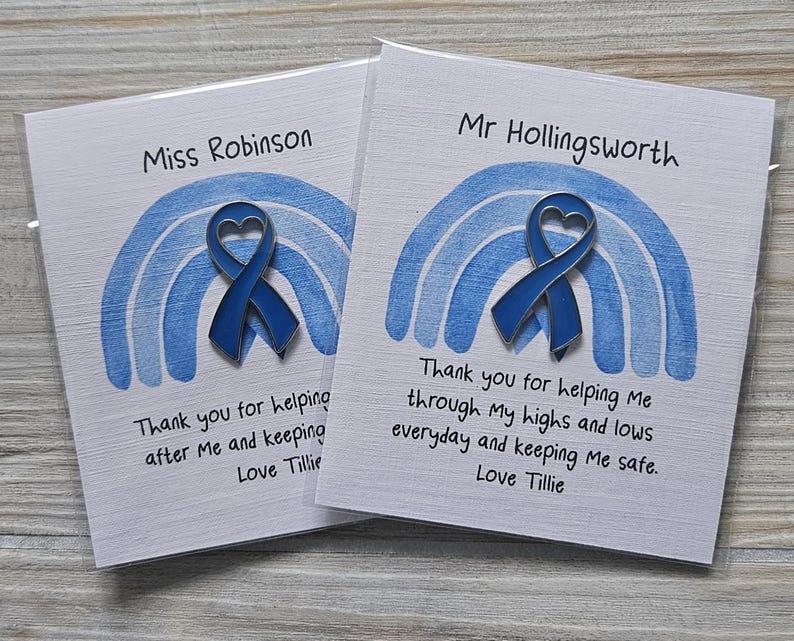 May include: Two white cards featuring a blue enamel heart-shaped ribbon pin centered over a blue painted rainbow graphic. The cards are personalized with names and printed text: Thank you for helping me through my highs and lows everyday and keeping me safe. Love Tillie.