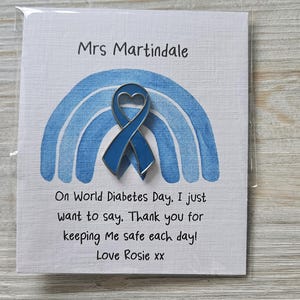 May include: A blue awareness ribbon pin with a heart-shaped cutout, centered on a white card featuring a blue watercolor rainbow. The card text reads: Mrs Martindale, On World Diabetes Day, I just want to say, Thank you for keeping Me safe each day! Love Rosie xx.