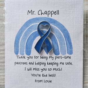 May include: A blue enamel awareness ribbon pin with a heart cutout, attached to a white card featuring a blue watercolor rainbow. The card text reads: Mr. Chappell, Thank you for being My part-time pancreas and helping keeping Me safe, I will Miss you so MUCH! You're the best! From Louie.