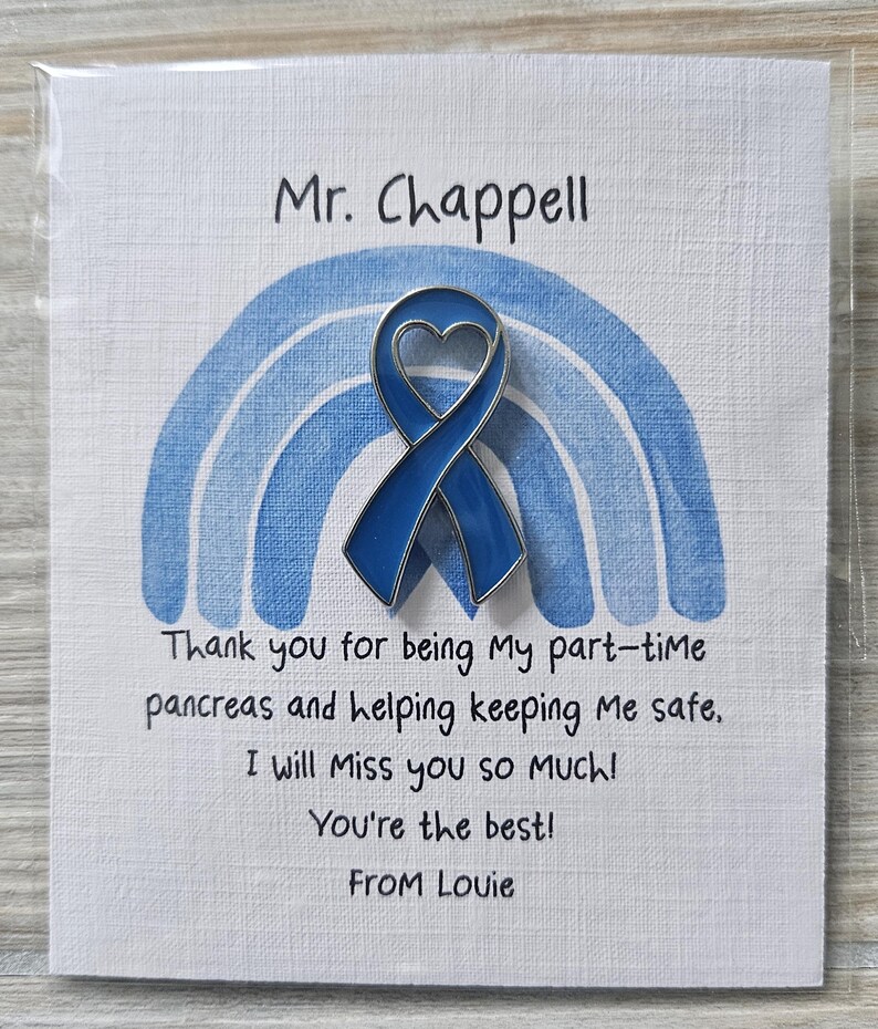 May include: A blue enamel awareness ribbon pin with a heart cutout, attached to a white card featuring a blue watercolor rainbow. The card text reads: Mr. Chappell, Thank you for being My part-time pancreas and helping keeping Me safe, I will Miss you so MUCH! You're the best! From Louie.