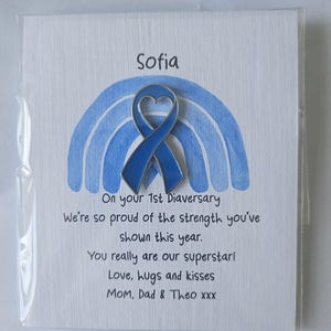 May include: A blue enamel awareness ribbon pin with a heart-shaped cutout, attached to a white card featuring a light blue watercolor rainbow. The card text reads: Sofia, On your 1st Diaversary, We're so proud of the strength you've shown this year.