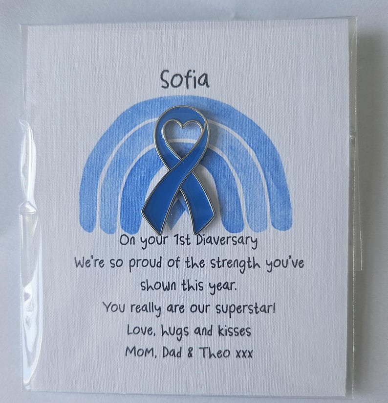May include: A blue enamel awareness ribbon pin with a heart-shaped cutout, attached to a white card featuring a light blue watercolor rainbow. The card text reads: Sofia, On your 1st Diaversary, We're so proud of the strength you've shown this year.